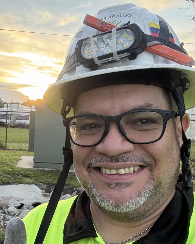 Greg Araujo Castillo smiling at sunrise on a job site, wearing a hard hat with a Venezuelan flag and a high-visibility shirt
